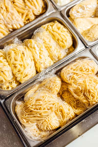 Freshly made Italian pasta varieties—including fettuccine, tagliatelle, and spaghetti—individually portioned and wrapped in plastic wrap, stored in stainless steel containers inside a professional kitchen prep station, showcasing authentic handmade pasta ready for cooking.