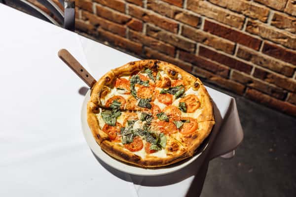 Fresh Margherita pizza with mozzarella, tomato slices, and basil served on a white plate atop a table with a brick wall backdrop.
