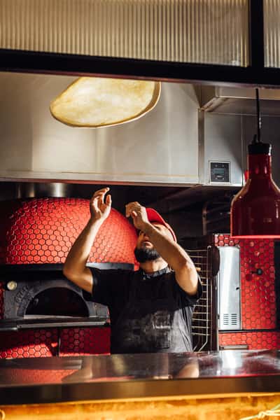 Chef tossing pizza dough high into the air in front of a blazing red-tiled wood-fired oven, capturing the artistry and tradition of authentic Italian pizza-making in a modern pizzeria setting.