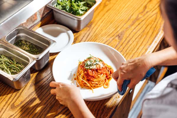 Chef preparing a classic spaghetti marinara dish topped with parmesan and basil in a professional kitchen with fresh herbs nearby.