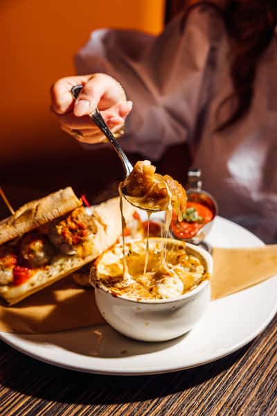 A hand lifting a spoonful of cheesy French onion soup from a white bowl, served with a meatball sub sandwich and marinara sauce on a white plate
