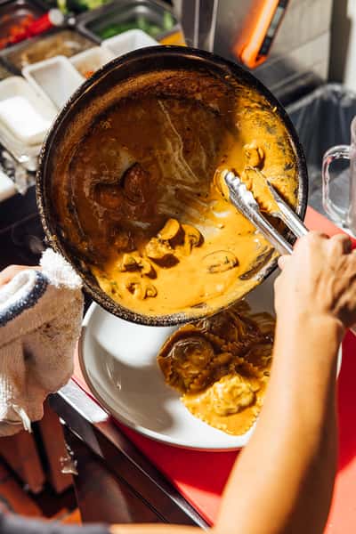 Rich mushroom cream sauce with sliced mushrooms being poured over stuffed ravioli from a sauté pan in a professional kitchen—ideal for highlighting handmade Italian pasta and behind-the-scenes culinary preparation.