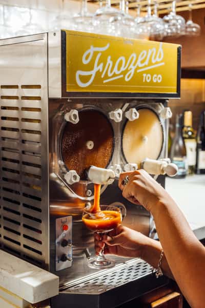 Bartender pouring a frozen cocktail from a dual-chamber slush machine labeled "Frozens To Go" into a coupe glass.
