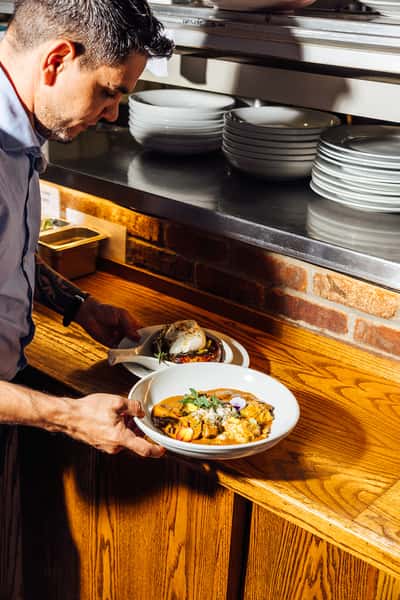 A restaurant server picking up two freshly plated dishes from a wooden counter—one featuring a sausage appetizer topped with burrata, the other a creamy pasta garnished with herbs and edible flowers.