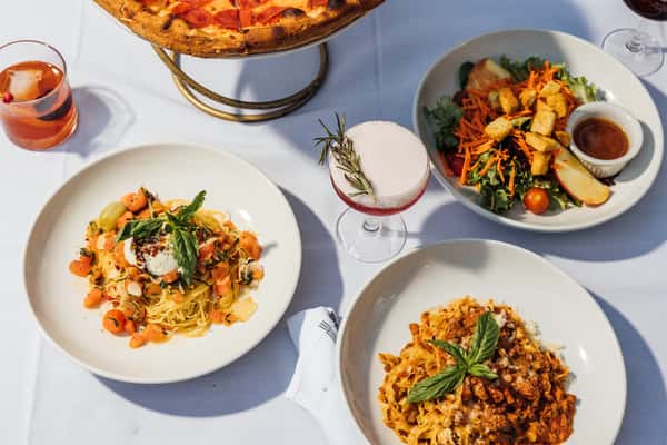 Overhead view of an Italian restaurant table featuring a pepperoni pizza on a stand, creamy burrata pasta with cherry tomatoes, hearty Bolognese tagliatelle, a fresh garden salad with apple and croutons, and a rosemary‑garnished craft cocktail on a white tablecloth—ideal for showcasing a complete Italian dining experience.