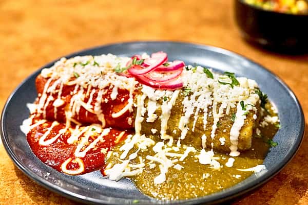 Enchiladas topped with red and green salsa, drizzled with crema and queso fresco, and garnished with pickled red onions and cilantro.