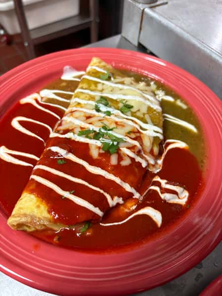 Colorful Mexican-style burrito smothered in red and green enchilada sauces, topped with melted cheese, drizzled crema, and chopped cilantro, served on a vibrant red plate at La Delicia Mexican Brunch & Cantina.