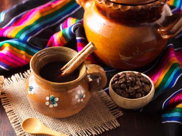 Traditional Mexican café de olla served in a clay mug with cinnamon stick, surrounded by coffee beans and colorful serape fabric.