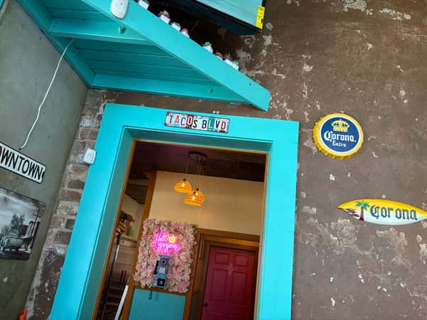 Vibrant entryway at La Delicia Mexican Brunch & Cantina featuring a turquoise door frame with a "TACOS BLVD" sign, rustic brick and stucco walls, Corona beer decor, and a floral wall with a neon "Hello Gorgeous" sign inside the cozy interior.