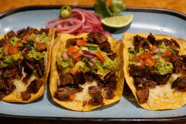 Three carne asada tacos topped with guacamole and pico de gallo, served on corn tortillas with melted cheese.