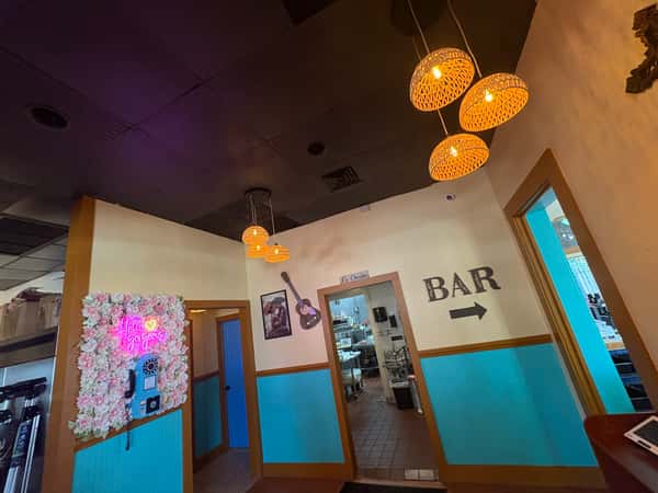 Colorful hallway inside a Mexican restaurant featuring woven pendant lights, a floral neon sign wall, and a sign pointing to the bar.