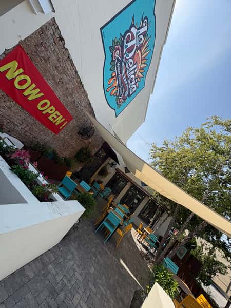 Colorful outdoor patio with teal and yellow seating, shaded by sail canopies, and a large La Delicia mural above a "Now Open!" banner.