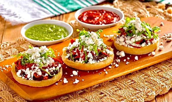 Trio of authentic Mexican sopes topped with refried beans, shredded lettuce, crumbled queso fresco, and salsa roja, served on a rustic wooden board with green and red salsas on the side.