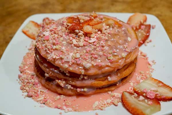 Stack of fluffy strawberry pancakes drenched in pink strawberry glaze, topped with crushed candy pieces and fresh strawberry slices for a sweet and colorful breakfast treat.
