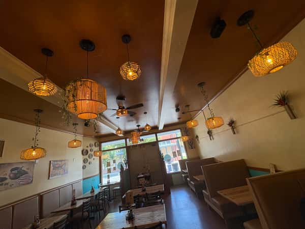 Warmly lit dining room with woven pendant lights, booth and table seating, wall art, and a view of the street through large windows.