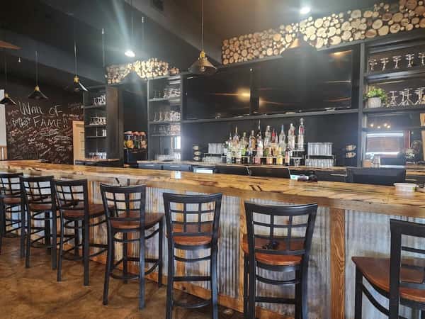 Rustic bar with wood and corrugated metal design, stocked shelves, a chalkboard "Wall of Fame," and high-top seating.