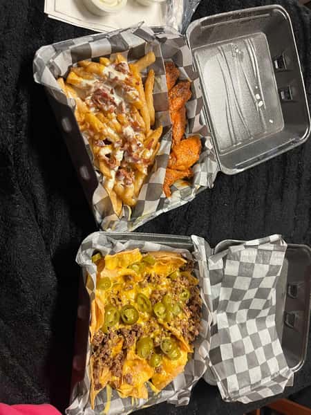 Loaded fries with bacon and ranch, buffalo wings, and beef nachos topped with cheese and jalapeños in black checkered paper trays.
