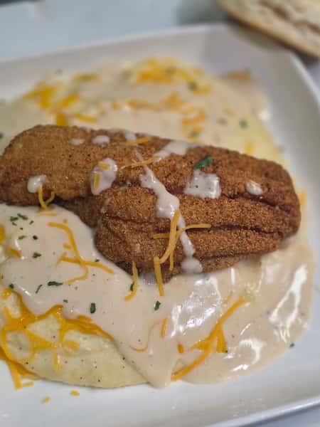 Fried catfish fillets served over creamy grits topped with shredded cheese and white gravy on a white plate.