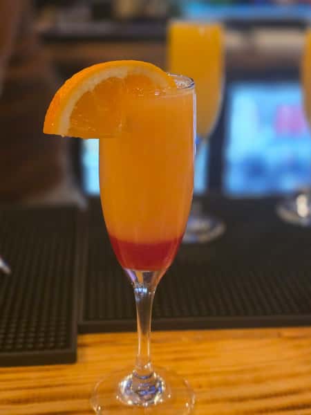 Tall champagne flute filled with a vibrant orange cocktail, layered over red syrup, and garnished with a fresh orange slice.