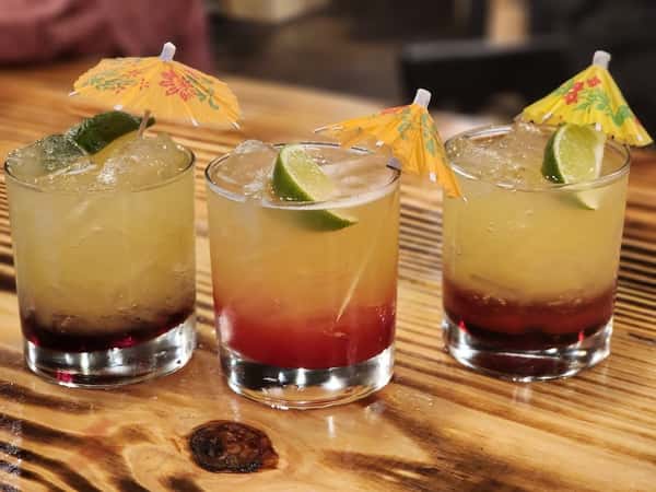 Three tropical cocktails garnished with lime wedges and colorful paper umbrellas, served over ice in short glasses on a wooden table.