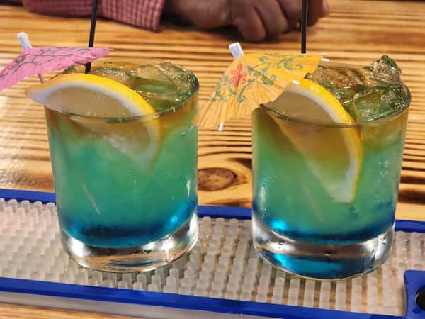 Two tropical blue cocktails with lemon slices and paper umbrellas served over ice on a bar counter.