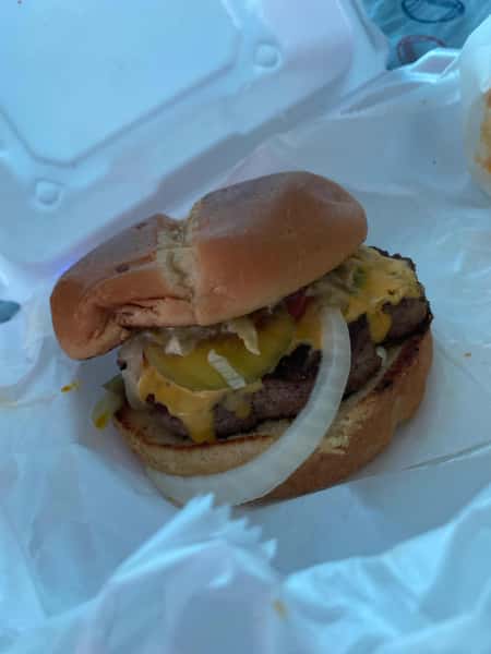 Cheeseburger with grilled beef patty, melted cheese, pickles, and raw onions on a toasted bun in a takeout container.