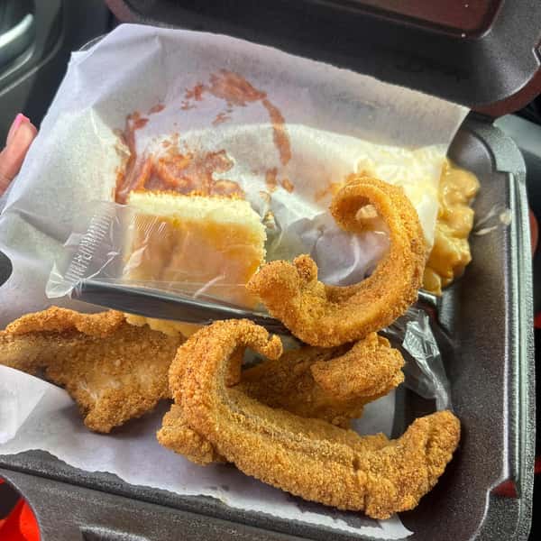 Fried catfish fillets with mac and cheese, a slice of cake in plastic wrap, and a side of spaghetti in a black takeout container.