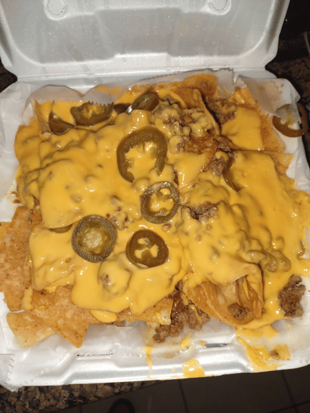 Loaded nachos topped with ground beef, creamy nacho cheese, and sliced jalapeños in a white foam takeout container.