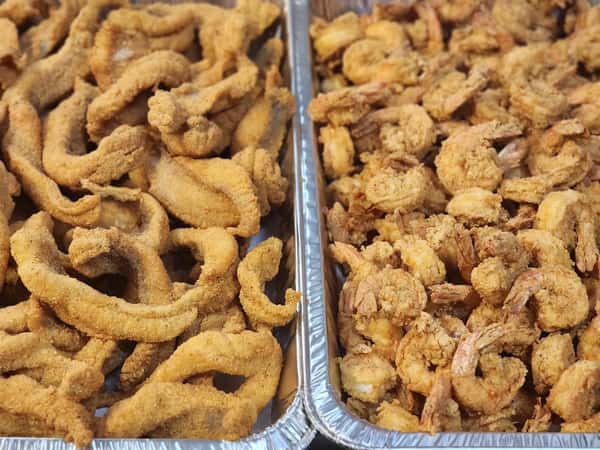 This image displays two large aluminum trays filled with crispy, golden-brown fried seafood. The tray on the left contains fried catfish strips, while the tray on the right is piled high with fried shrimp, all coated in a seasoned breading. Perfect for a Southern-style seafood feast or catering event.