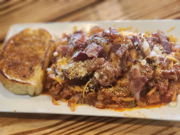 This image shows a hearty plate of spaghetti topped with a rich meat sauce, crumbled bacon, shredded cheese, and a sprinkle of grated parmesan. It's served with a slice of golden-brown garlic toast on the side.
