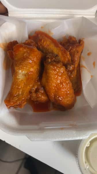 Buffalo-style chicken wings coated in hot sauce, served in a white foam takeout container with a side of ranch dressing.