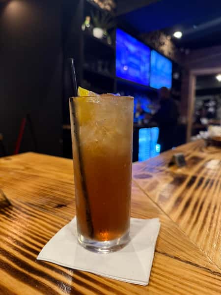 This appears to be a tall cocktail—possibly a Long Island Iced Tea—served over ice in a highball glass, garnished with a lemon wedge, and accompanied by a straw. It's placed on a napkin atop a wooden bar counter.