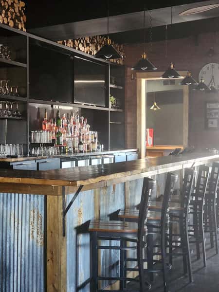 Rustic bar setup with corrugated metal paneling, high wooden stools, hanging pendant lights, and a variety of liquor bottles on display.