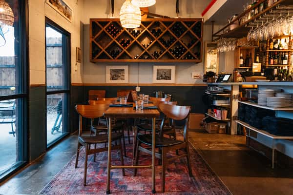 Warmly lit interior view of The Downing Bottles and Bites, featuring a central wooden dining table, large diamond wine rack, and service bar.
