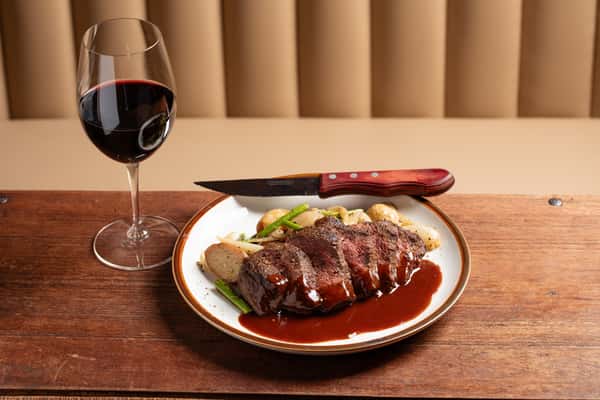 Sliced steak with rich sauce and vegetables, served with red wine on a dark wood table at The Downing Bottles and Bites.