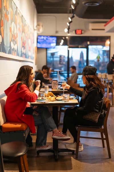 Interior view of The Downing Bottles & Bites showing two women, one in red, toasting with champagne and sharing a cheese board next to a colorful floral wall mural.
