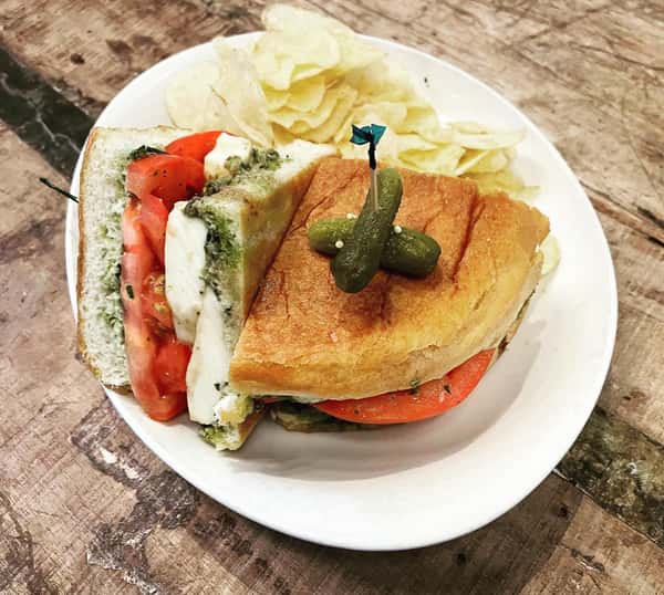 Pesto Caprese sandwich featuring mozzarella and tomato on toasted bread, served with crisp chips and pickles at The Downing Bottles and Bites.