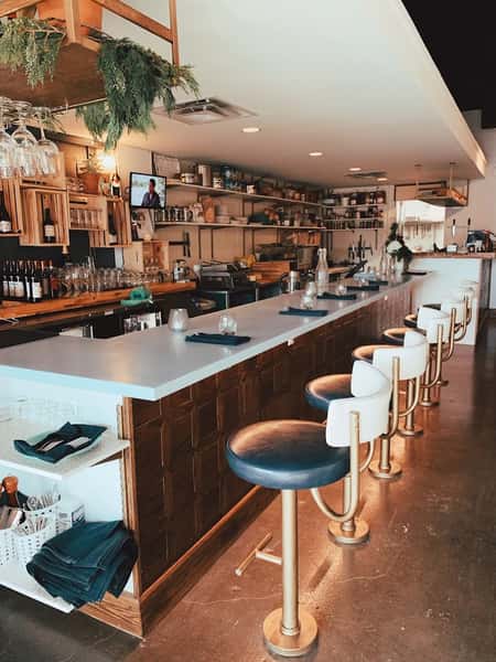 Modern interior bar scene at The Downing Bottles & Bites featuring navy and brass bar stools, a white counter, and industrial open shelving.