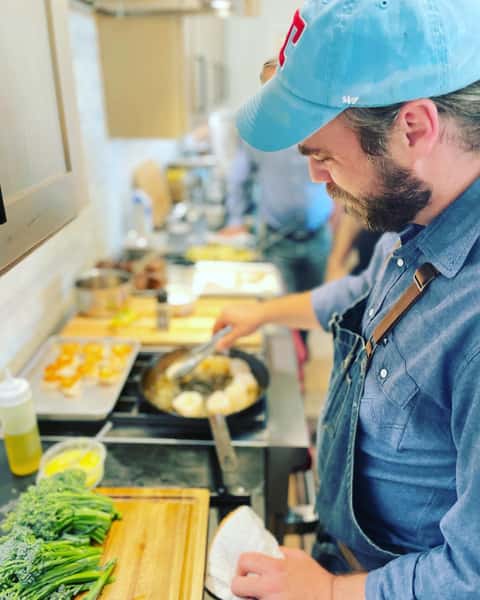 Bearded chef in a blue apron searing scallops and preparing broccolini in the kitchen at The Downing Bottles and Bites.