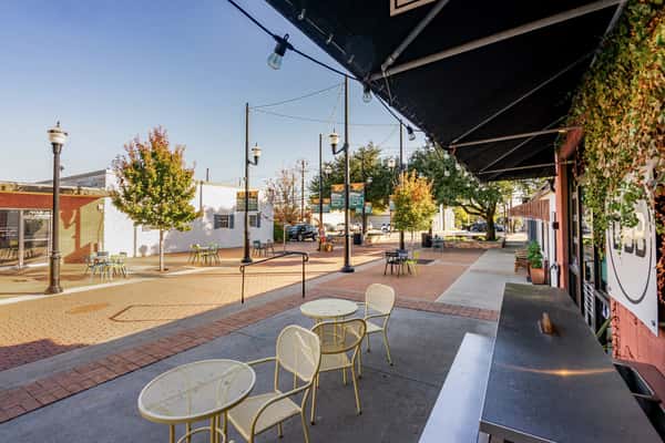 The Downing Bottles and Bites sunny outdoor patio featuring yellow bistro tables, a black awning, and a wide brick pedestrian plaza with string lights.