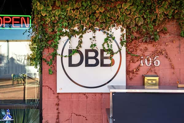Exterior sign for The Downing Bottles and Bites (DBB) draped in ivy against a pink brick wall, featuring a neon "OPEN" sign.