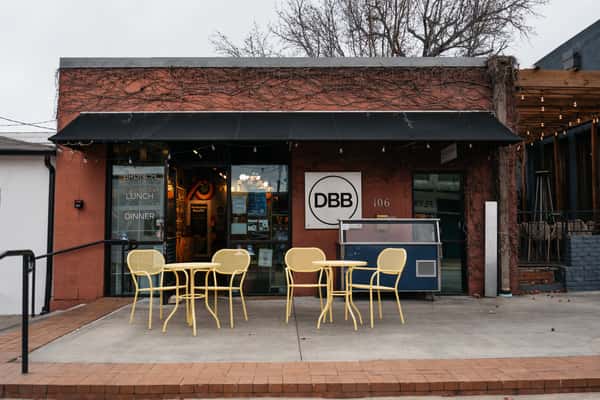 Exterior view of The Downing Bottles & Bites (DBB) featuring a red brick facade, black awning, and bright yellow sidewalk patio furniture.
