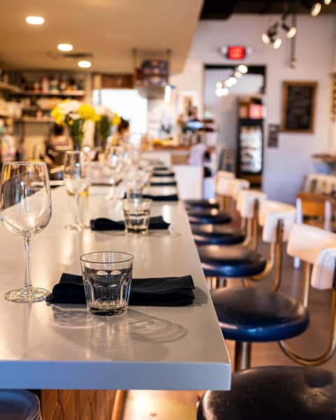 Warm interior shot of The Downing Bottles and Bites bar, highlighting the gray counter, wine glasses, and row of navy blue bar stools.
