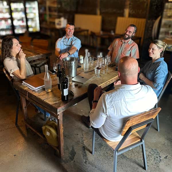 Interior shot of diverse patrons laughing during a red wine tasting at a rustic wooden table in The Downing Bottles and Bites.