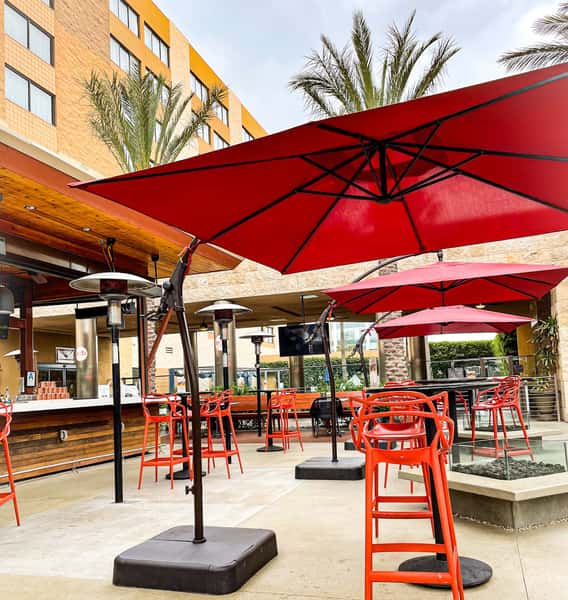 Modern outdoor patio at edb (eat. drink. be.) featuring red umbrellas, high-top seating, fire pit tables, and an open-air bar surrounded by palm trees.