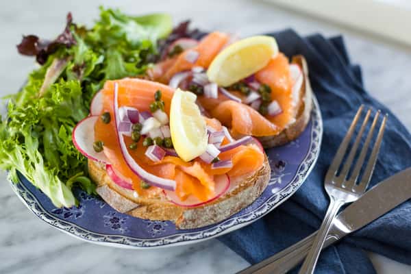 A vibrant open-faced sandwich is beautifully arranged on a patterned plate, topped with silky slices of smoked salmon, delicate ribbons of fresh vegetables, and thin lemon slices. Garnished with herbs and colorful edible flowers, the dish feels fresh, elegant, and full of bright, refined flavors—perfectly capturing a light and sophisticated café experience.