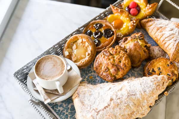 A cozy café-style spread featuring a cappuccino with latte art, surrounded by an assortment of freshly baked pastries. The selection includes croissants, buns, and filled pastries, all golden and inviting, presented on elegant patterned plates for a warm, artisanal feel.