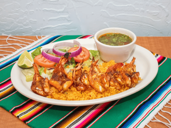 Grilled shrimp plate with Mexican rice, side salad, lime wedge, and a bowl of caldo or soup, served on a colorful Mexican tablecloth.