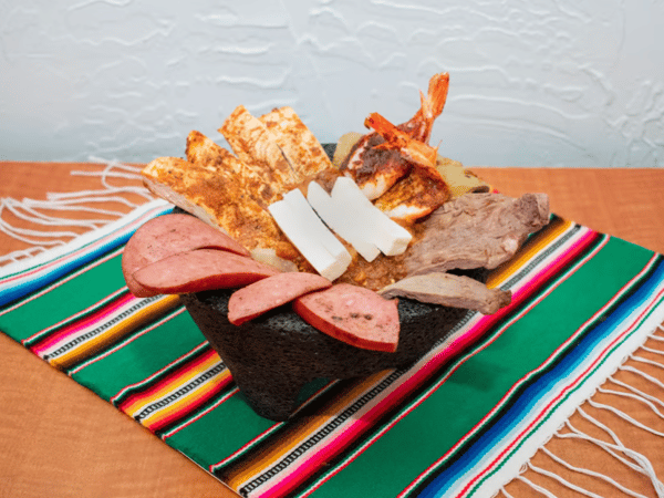 Molcajete platter filled with grilled steak, shrimp, chicken, cactus, sausage, and queso fresco, served sizzling hot in a traditional stone bowl atop a colorful Mexican table runner.