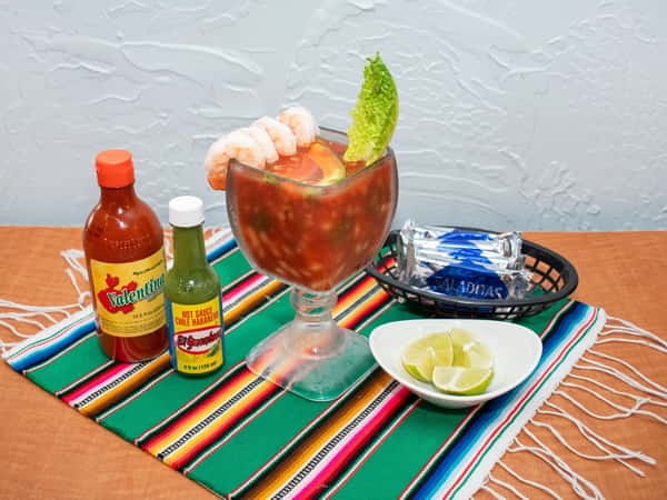 Mexican shrimp cocktail served in a large goblet with whole shrimp, fresh lime, and lettuce, accompanied by Valentina and El Yucateco hot sauces, saltine crackers, and lime wedges on a traditional sarape.